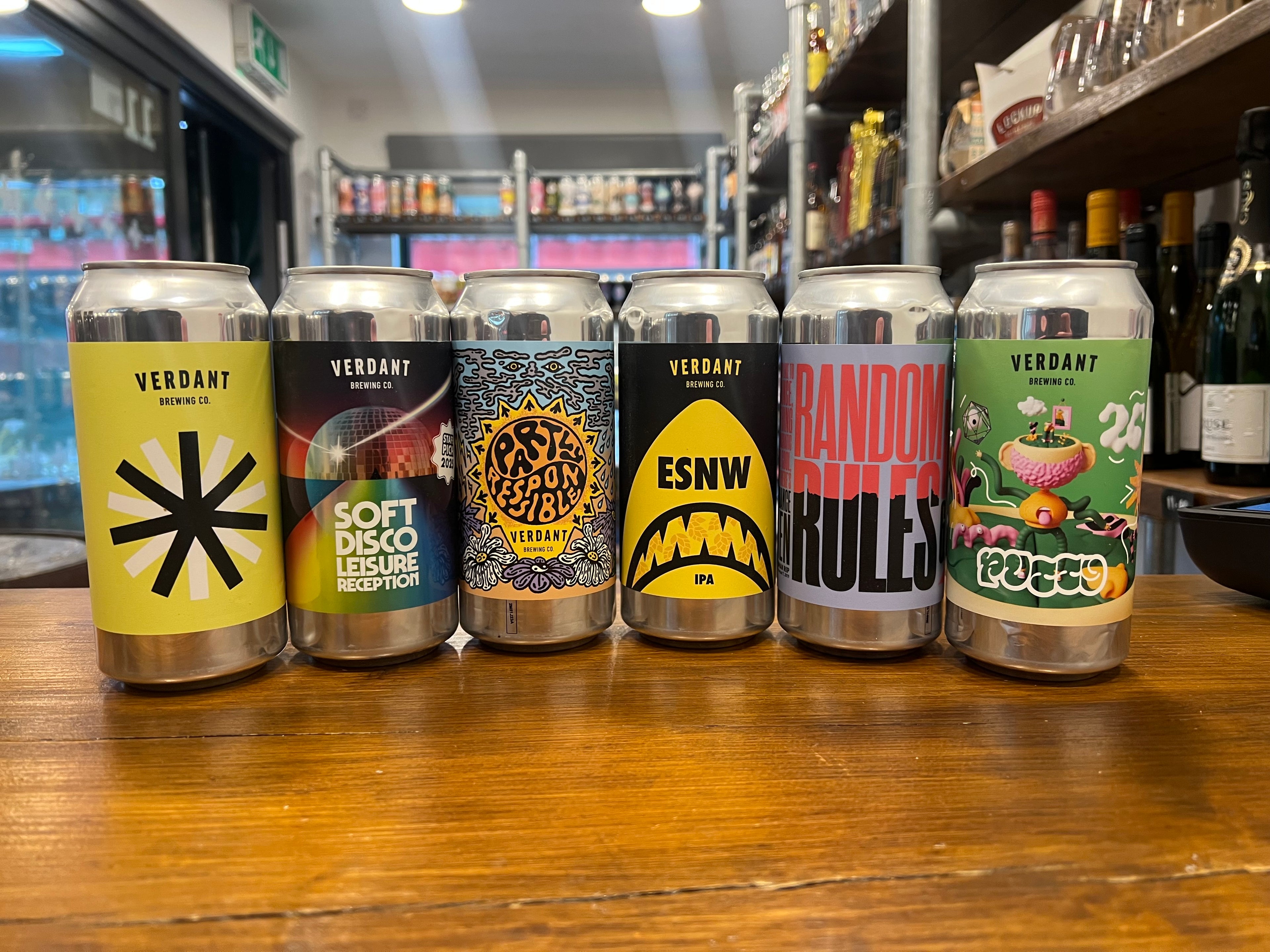 Six Verdant beer cans with various designs on a wooden surface in a bar setting.